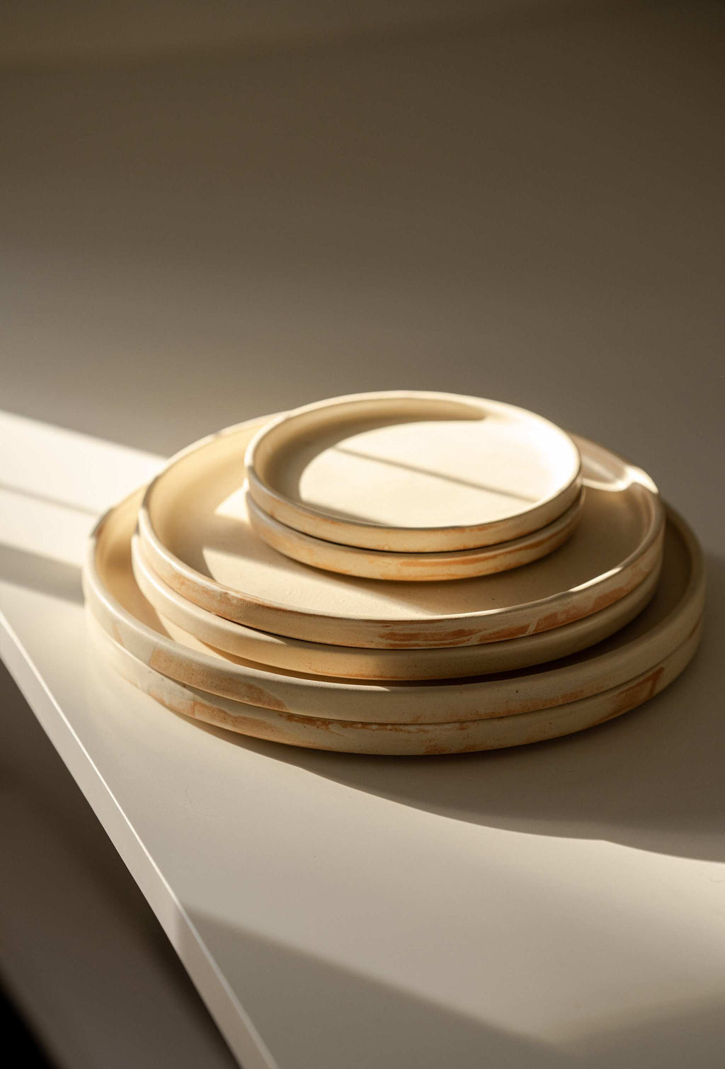 Stack of six beige matte stoneware plates in three sizes on a white surface in warm slanting sunlight