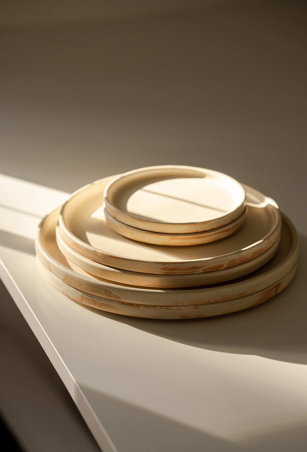 Stack of six beige matte stoneware plates in three sizes on a white surface in warm slanting sunlight
