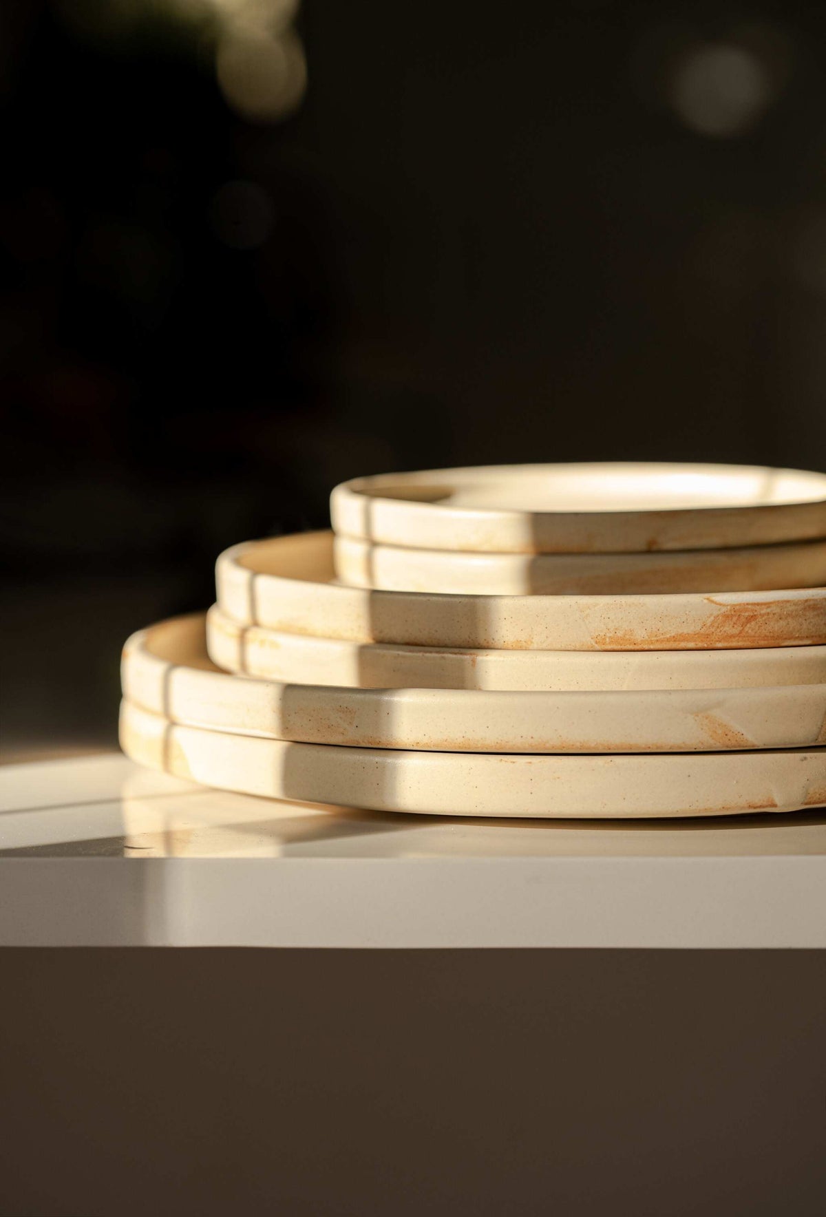 Side profile of six stacked beige matte stoneware plates with rust markings against a dark background