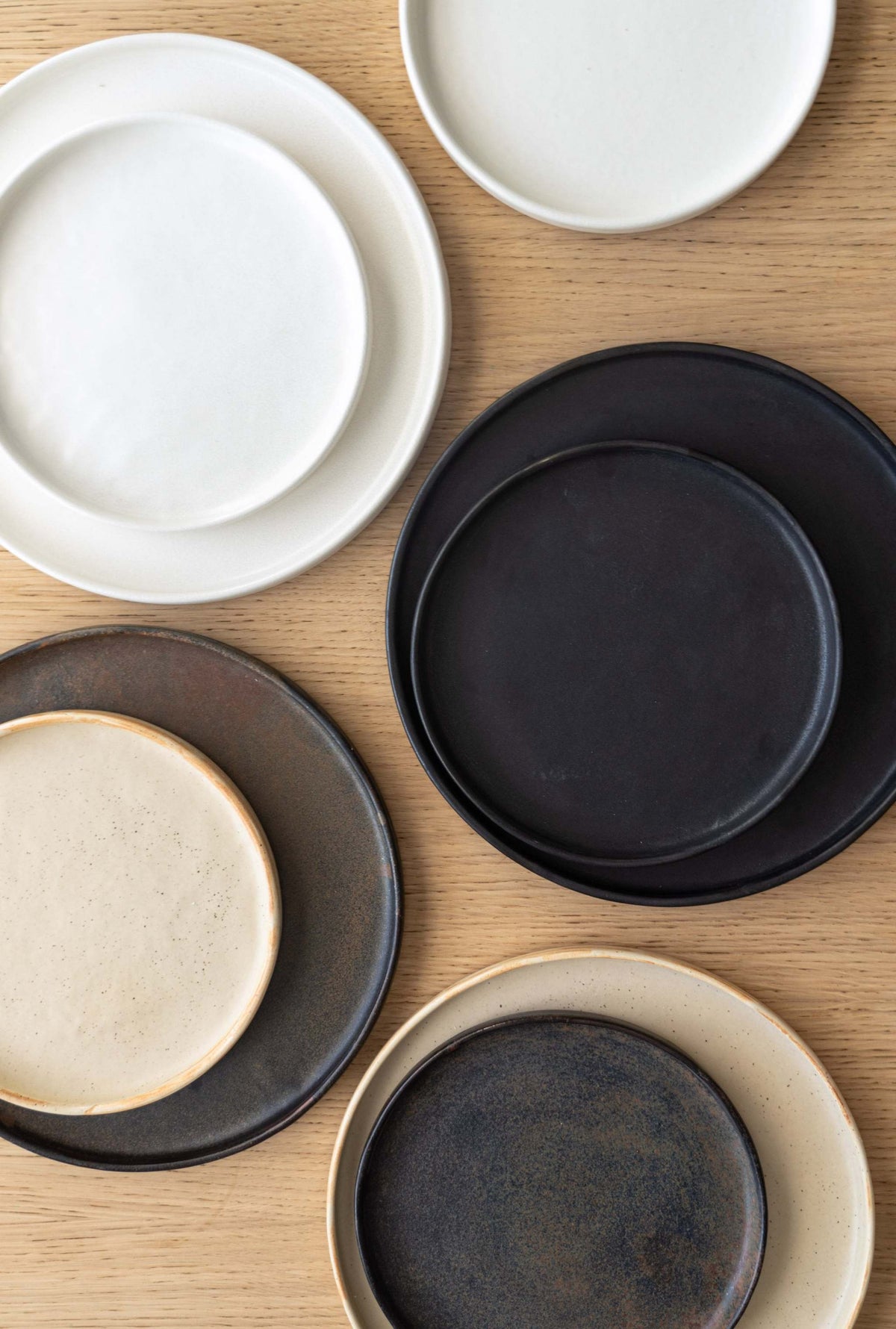 Matte stoneware plates in white, black, bronze and speckled beige paired in two sizes on oak, from above