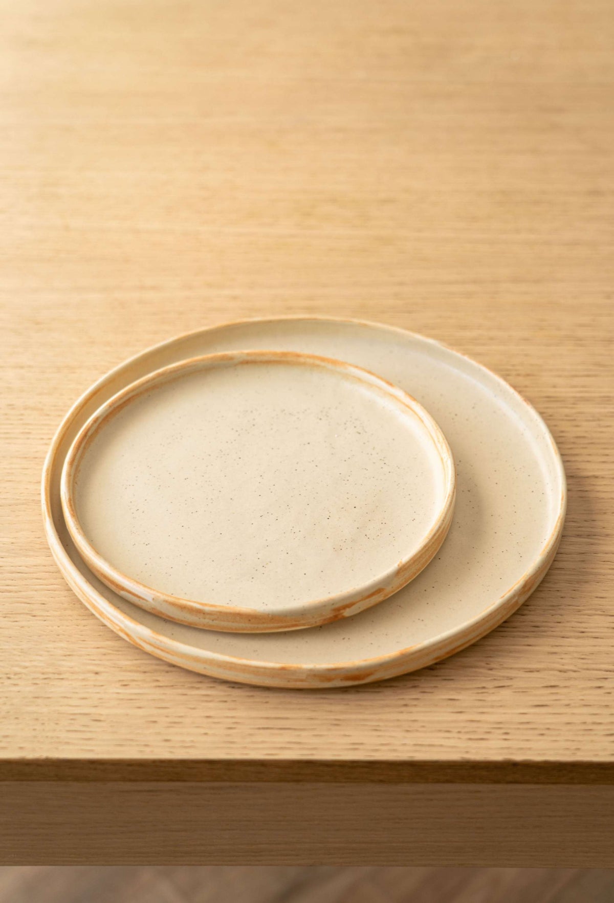 Two beige matte stoneware plates with rust brushed rims, the smaller nested in the larger on an oak table