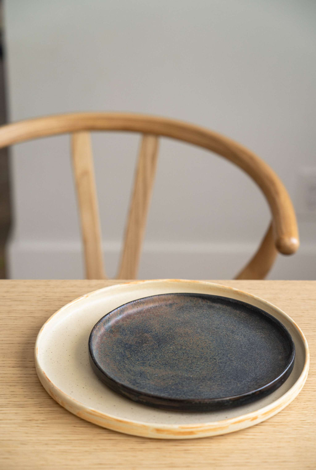Beige matte stoneware dinner plate under a smaller dark bronze plate on an oak table with a wooden chair behind