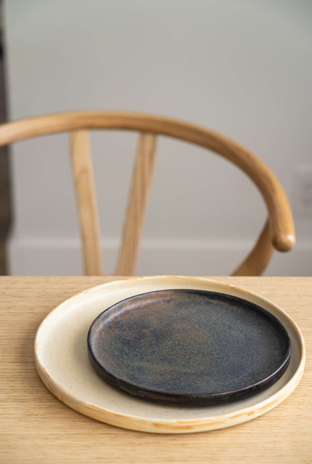 Beige matte stoneware dinner plate under a smaller dark bronze plate on an oak table with a wooden chair behind