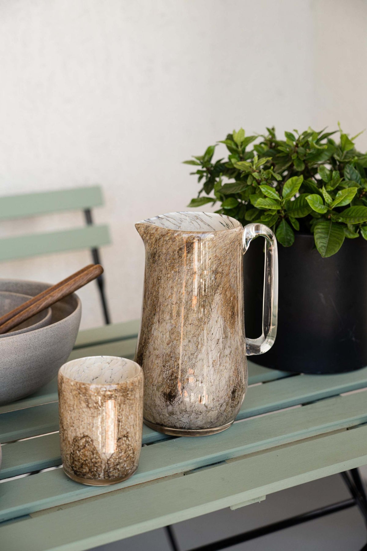 Marbled glass pitcher and glass cups on a pale green outdoor table beside a potted green plant