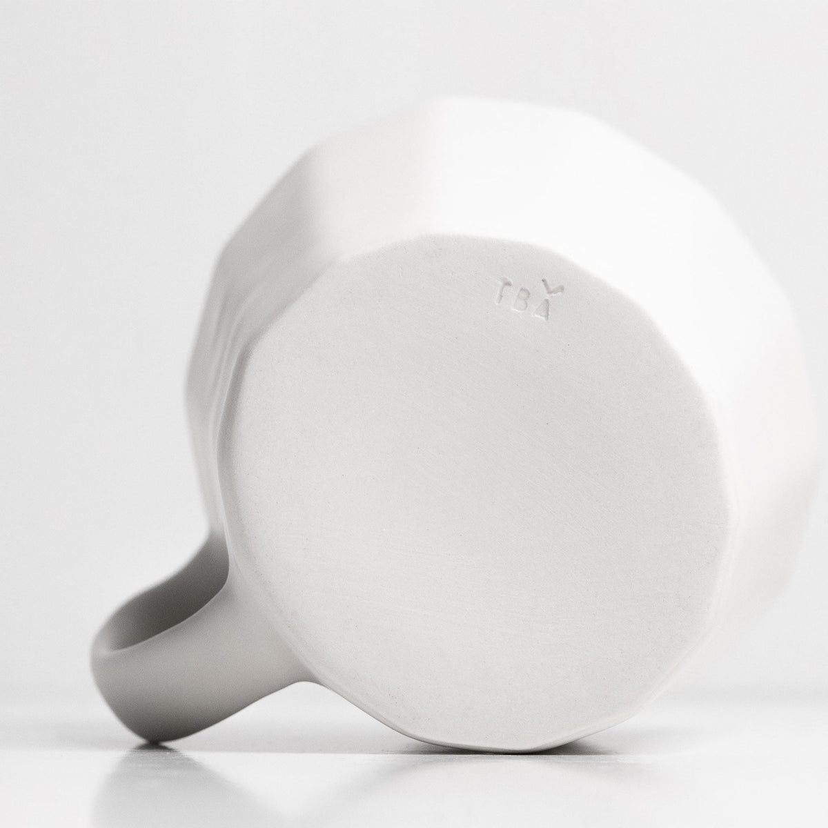 Underside of a white porcelain coffee mug showing the faceted walls, handle and unglazed foot