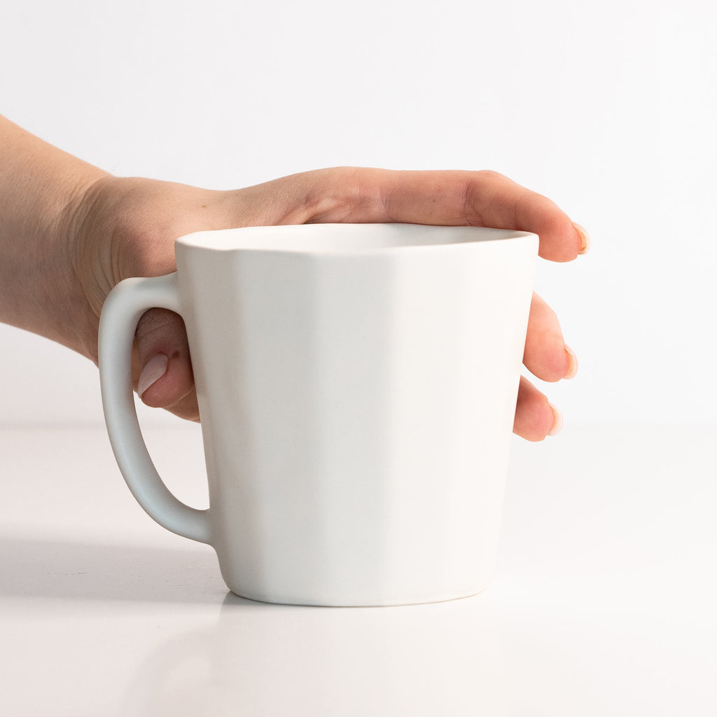 Hand resting on a Silk White porcelain coffee mug to show its size and softly faceted oval body