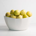 Large faceted porcelain serving bowl in Silk White piled with green apples against a pale studio backdrop