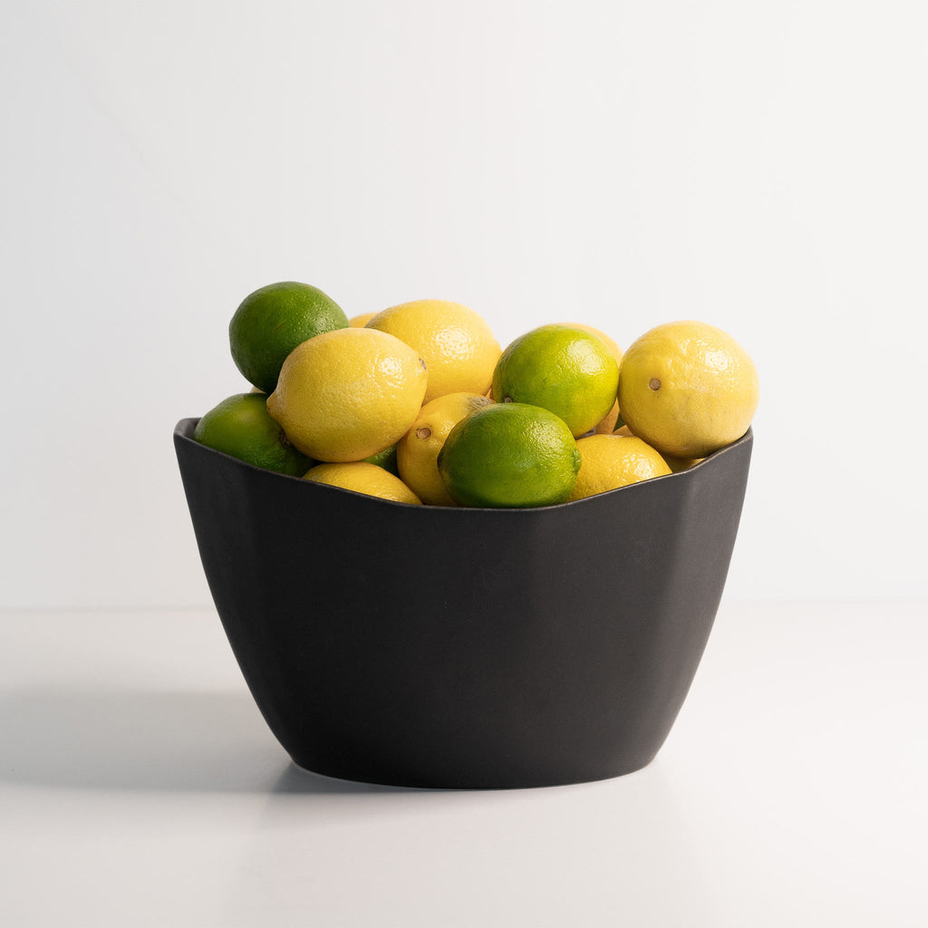 Medium faceted porcelain bowl in Mica Black filled with lemons and limes on a pale studio surface