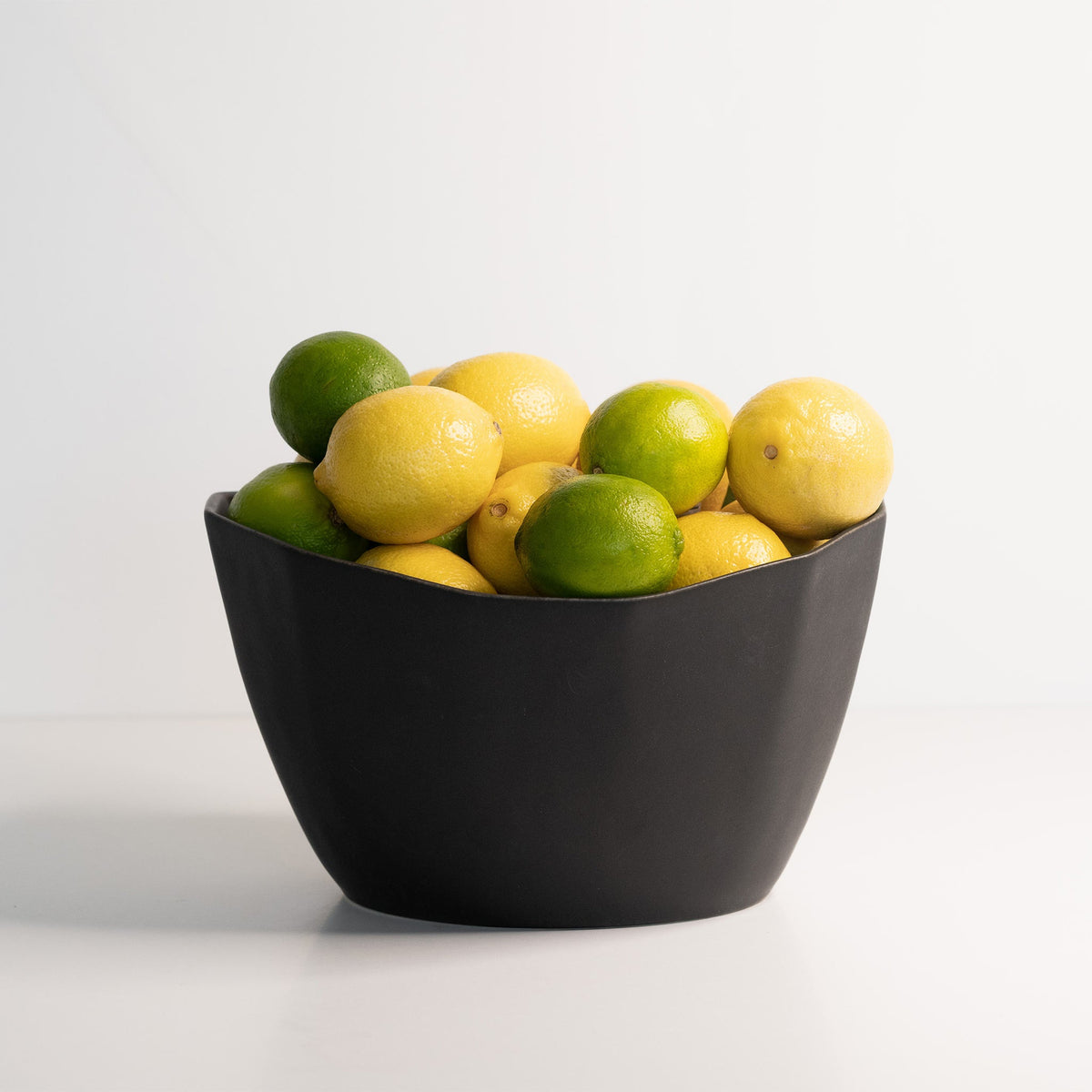 Medium faceted porcelain bowl in Mica Black filled with lemons and limes on a pale studio surface