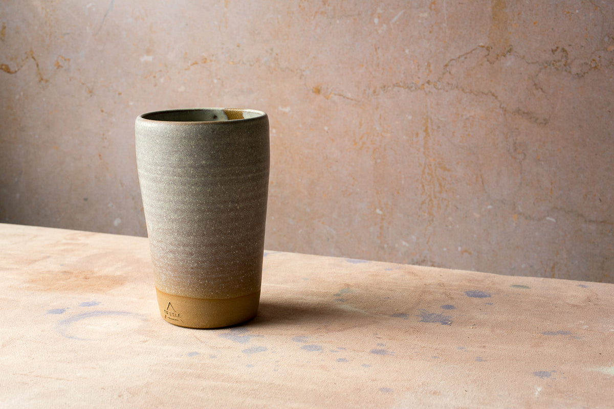 Tall hand-thrown stoneware cup in a speckled oat glaze with a bare clay foot on a sandstone-colored surface