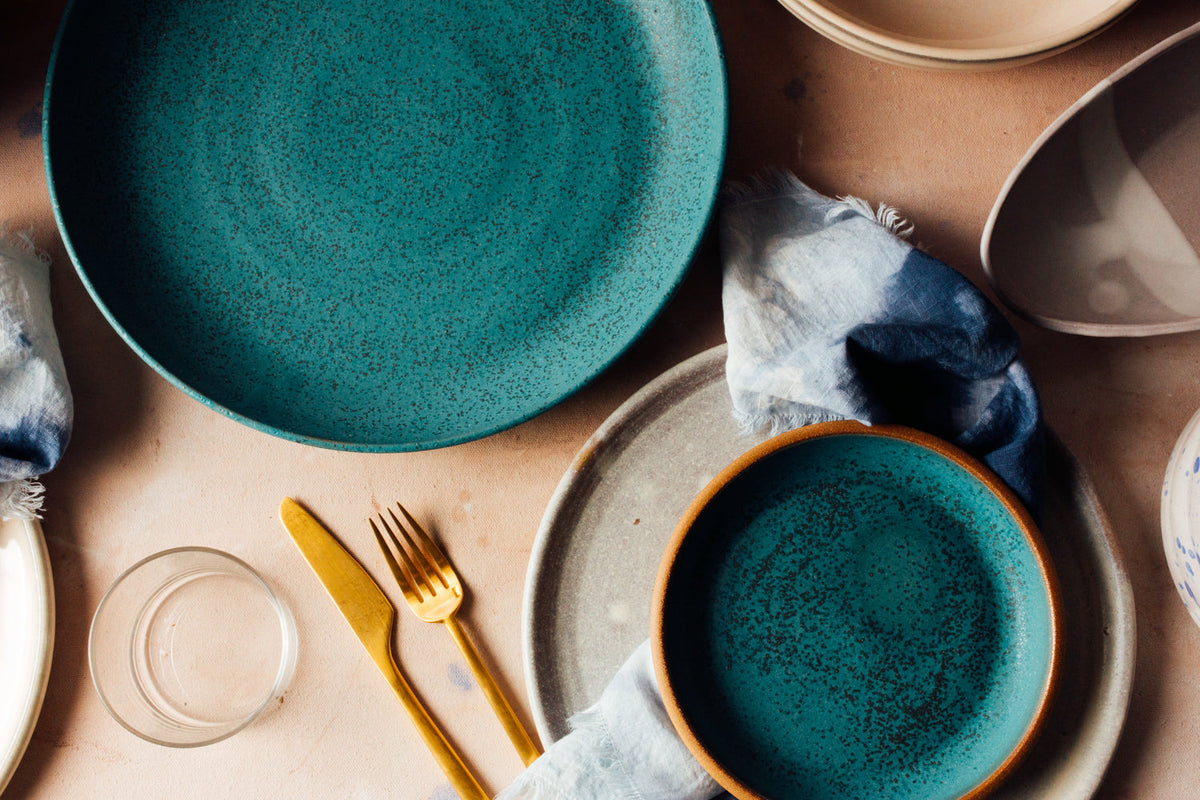 Overhead table setting with Fat Rim stoneware plates in speckled turquoise, brass flatware and indigo napkins
