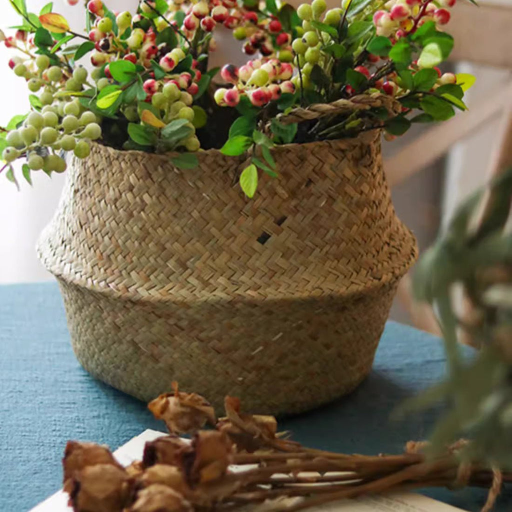 Woven seagrass belly basket holding a bunch of berry branches on a blue cloth, with dried roses below