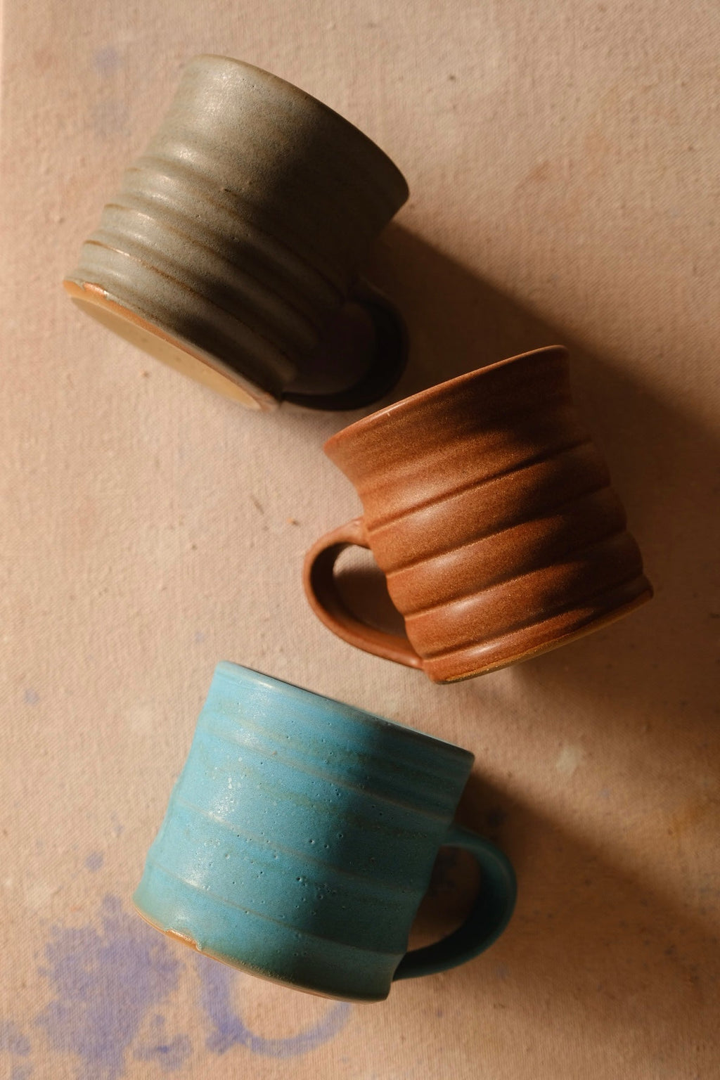 Three ridged hand-thrown stoneware mugs in grey-green, rust red and turquoise glazes on a sand-colored surface