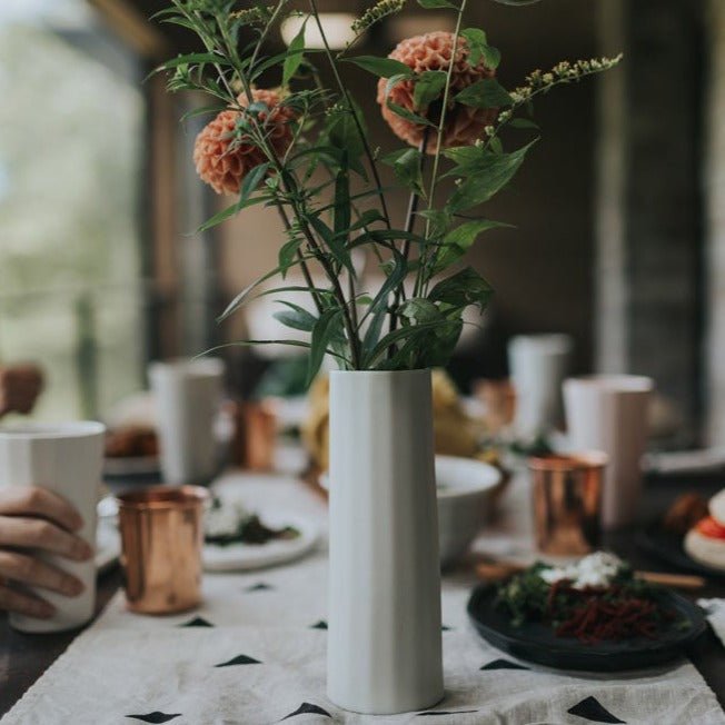 White porcelain bud vase of peach and green flowers on a set dinner table with copper mugs