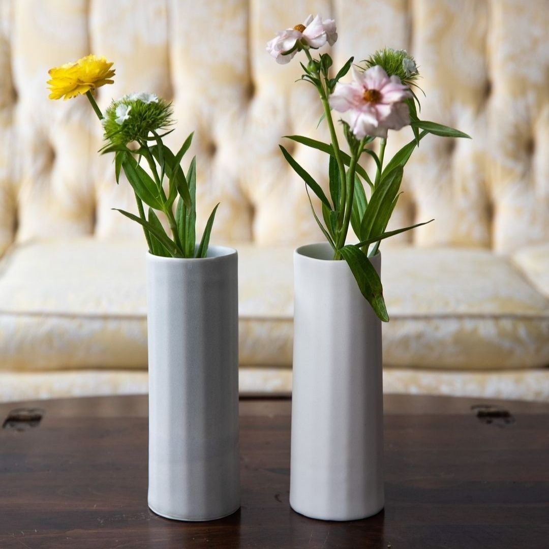 Two white porcelain bud vases of wildflowers on a dark wood table in front of a cream sofa