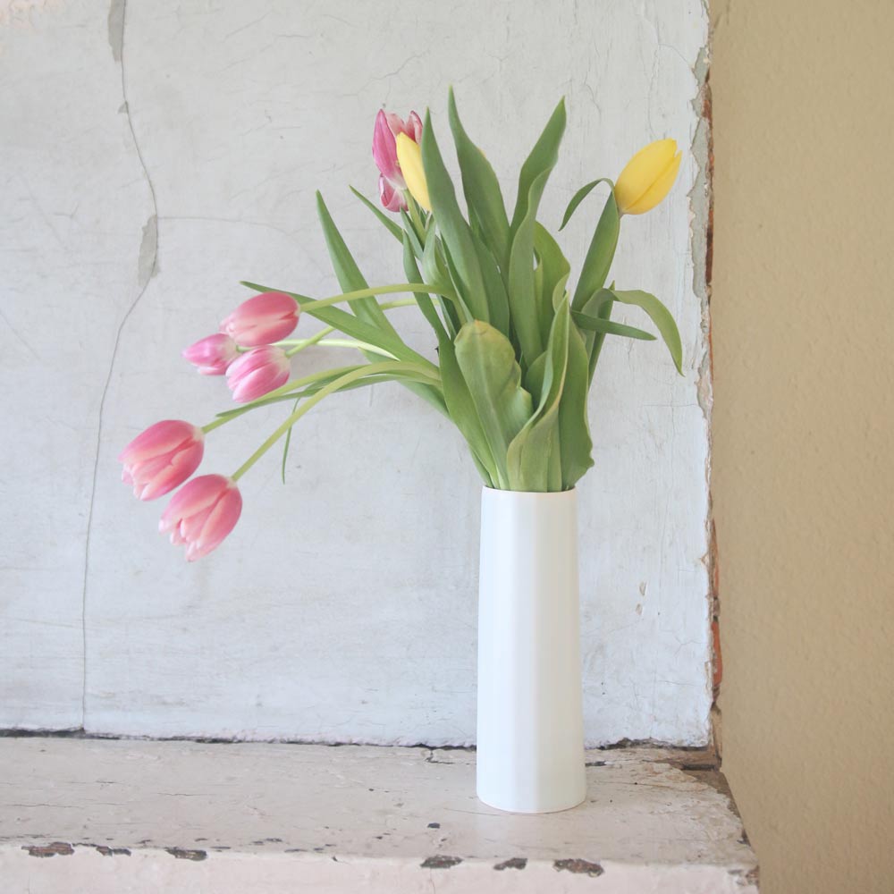 Slim white porcelain bud vase of pink and yellow tulips on a whitewashed ledge in soft daylight