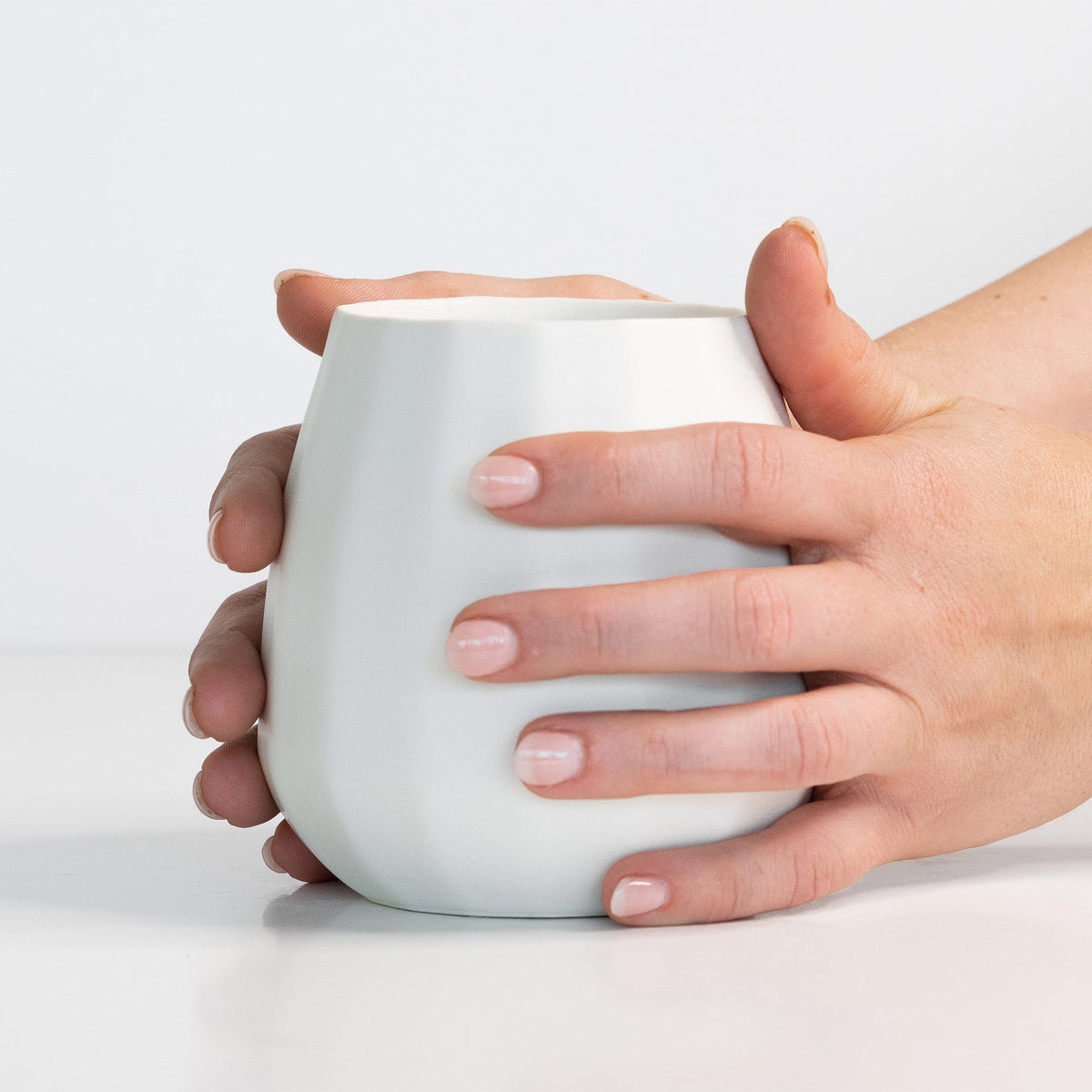 Hands holding a Silk White porcelain stemless wine glass, its rounded faceted body filling the palm