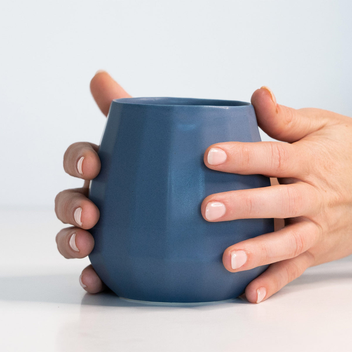Two hands holding a Pisgah Blue porcelain stemless wine glass to show its size against the palm