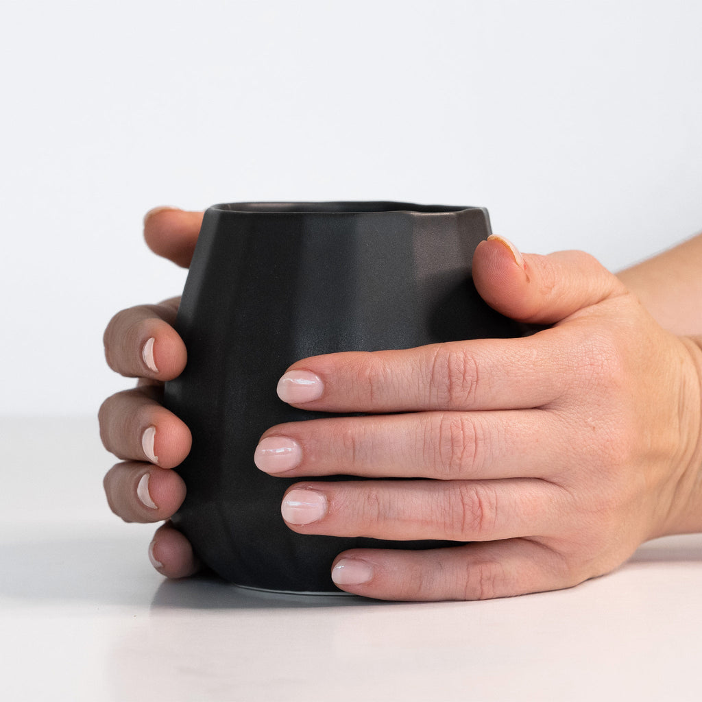 Two hands cupping a Mica Black porcelain stemless wine glass to show its palm-sized faceted shape