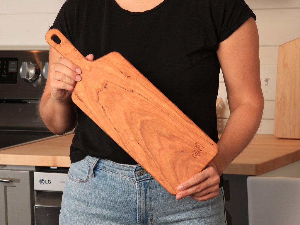Woman in a black tee holding a narrow cherry paddle serving board at her hip in a kitchen
