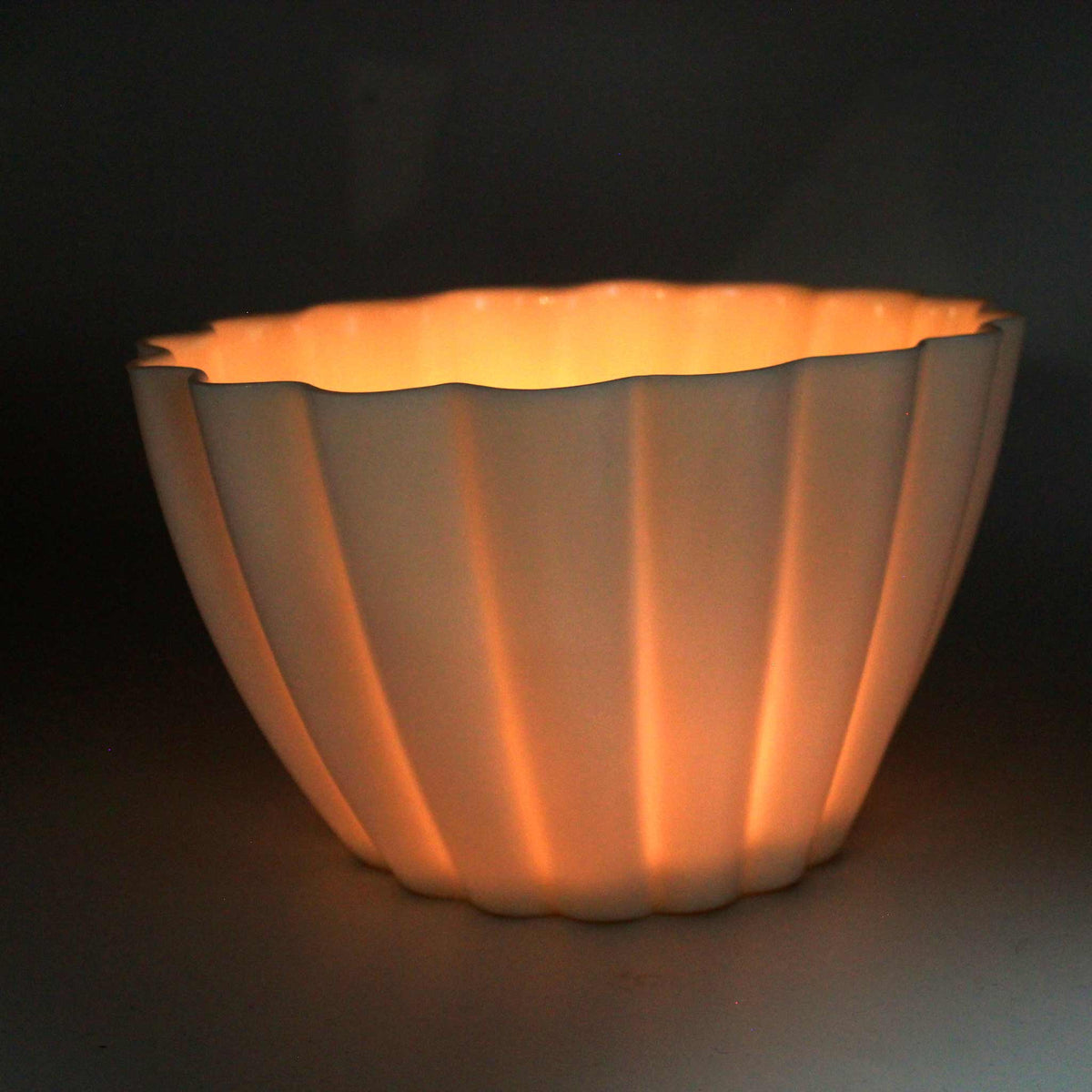 Fluted porcelain candle holder lit in a dark room, candlelight glowing through its translucent ribs