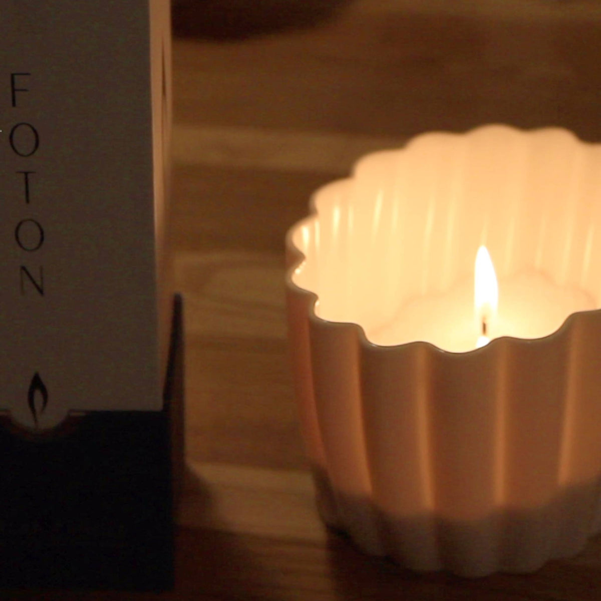 Lit fluted porcelain candle holder glowing warm on a wood table, the flame rising above its scalloped rim