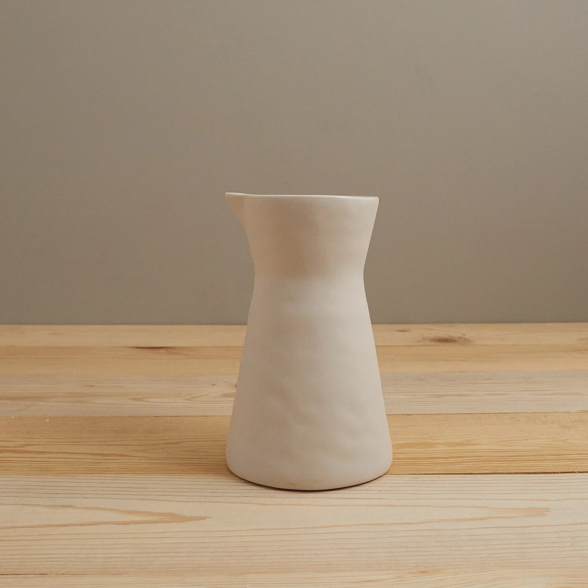 Hand-shaped matte pearl stoneware carafe standing on a light wood table against a warm taupe wall
