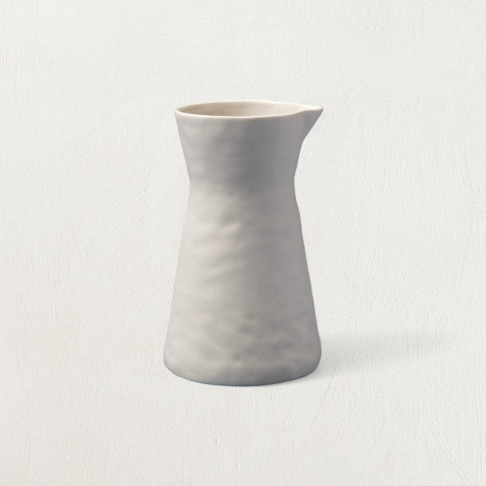 Matte pearl stoneware carafe with a gently dimpled, hand-shaped surface and pinched spout on a pale backdrop