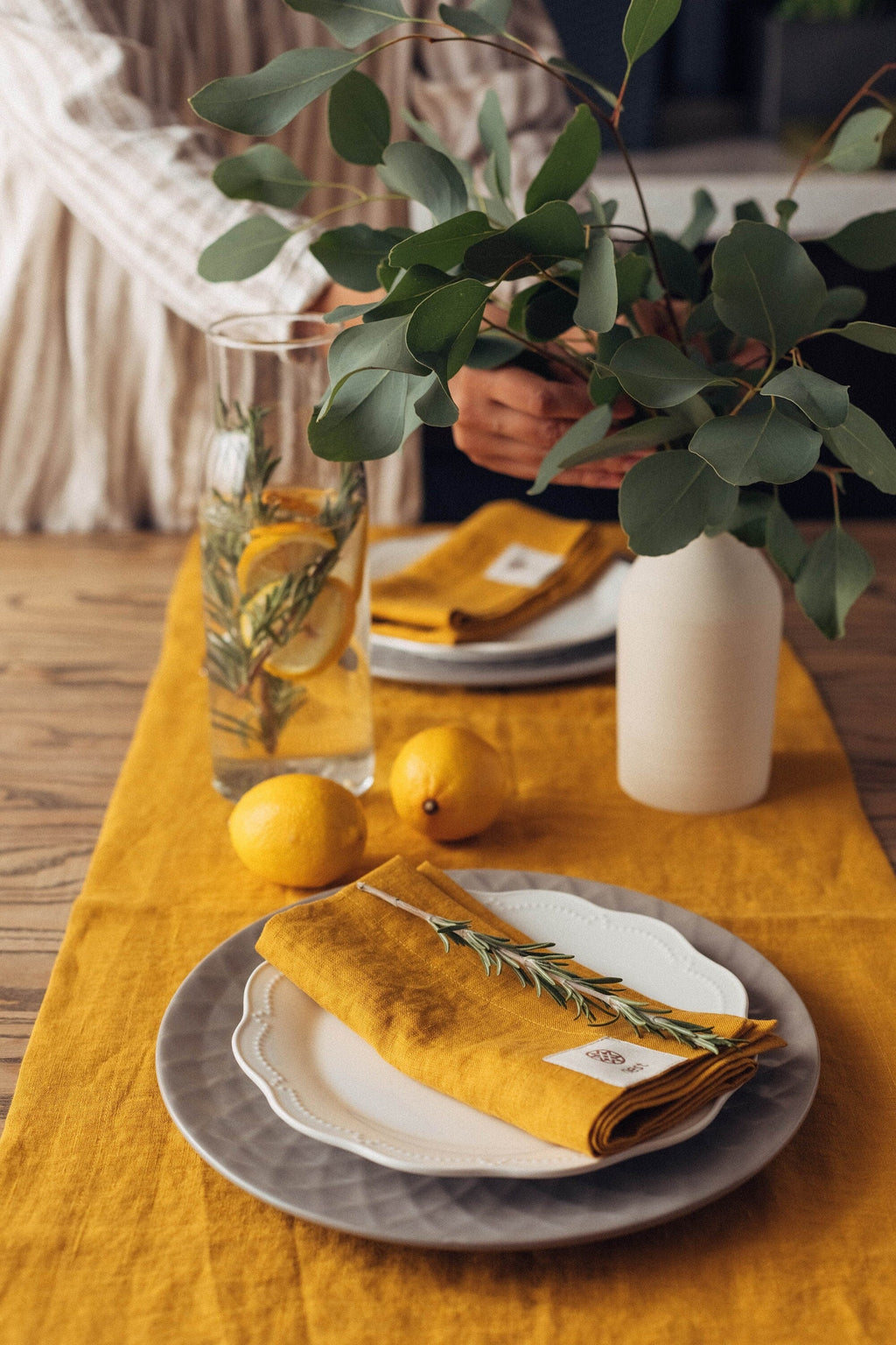 Golden yellow linen runner with a matching napkin, a rosemary sprig, lemons and a vase of eucalyptus