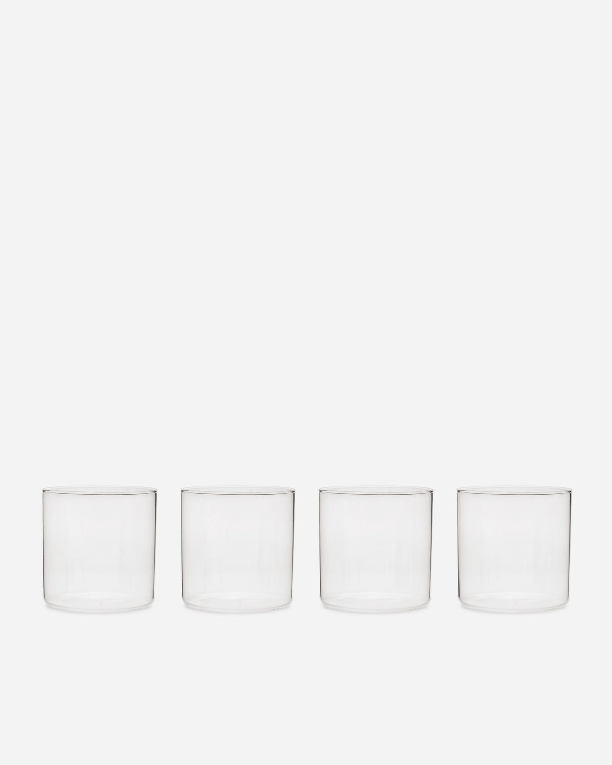 Four short handblown glass tumblers with straight sides and thin rims lined up on a pale grey background