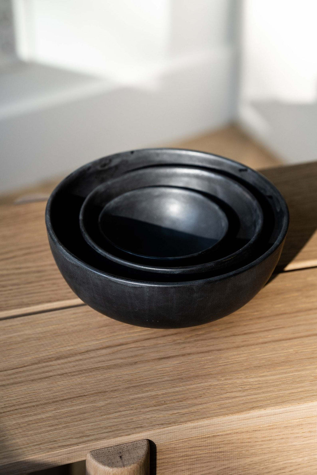 Three matte black stoneware bowls nested together on a light wood surface beside a sunny wall