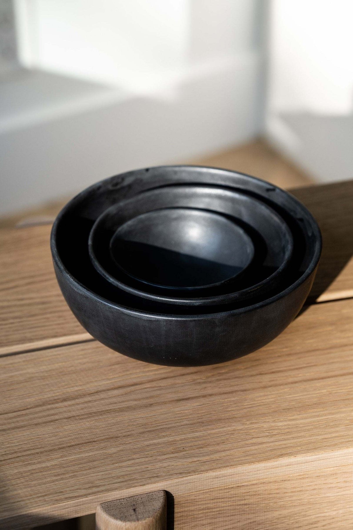 Three matte black stoneware bowls nested together on a light wood surface beside a sunny wall
