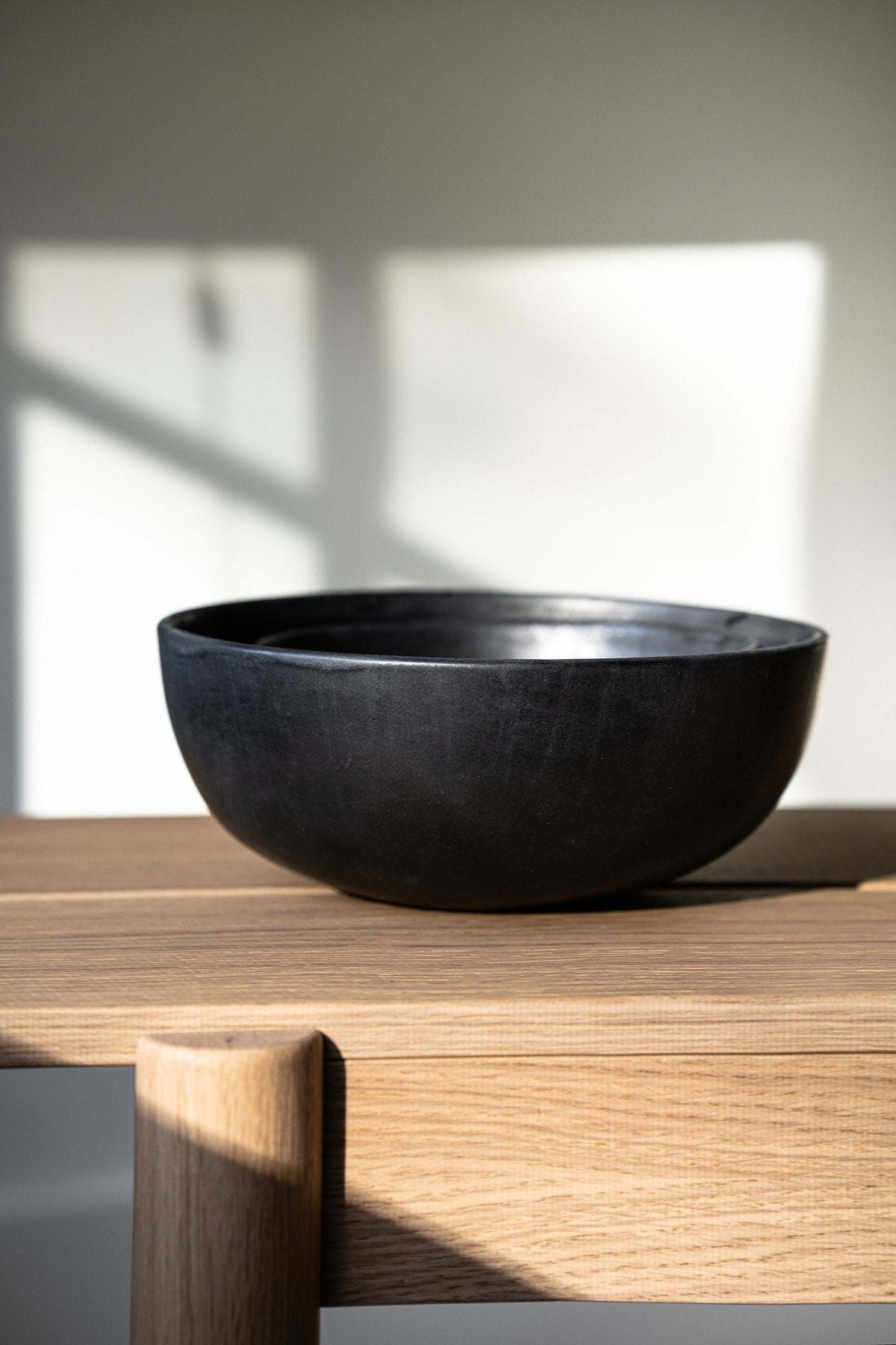 Matte black stoneware serving bowl on a pale wood table in soft window light with a long shadow
