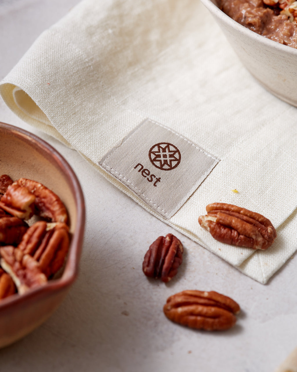 Ivory linen napkin with a woven maker's label beside bowls of pecans on a pale stone counter top