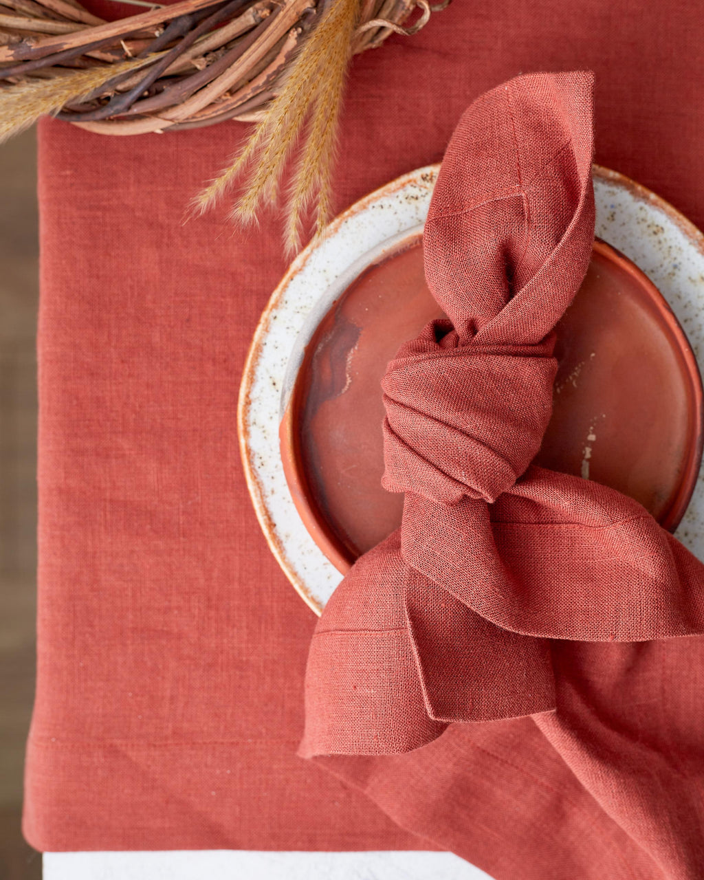 Russet linen runner with a knotted matching napkin on a speckled stoneware plate beside dried grass