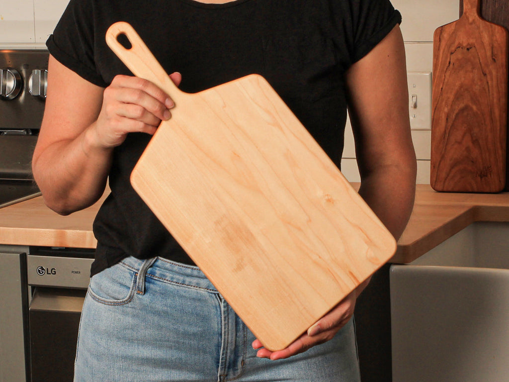 Woman in a black tee holding a wide maple paddle serving board in a kitchen, walnut board behind