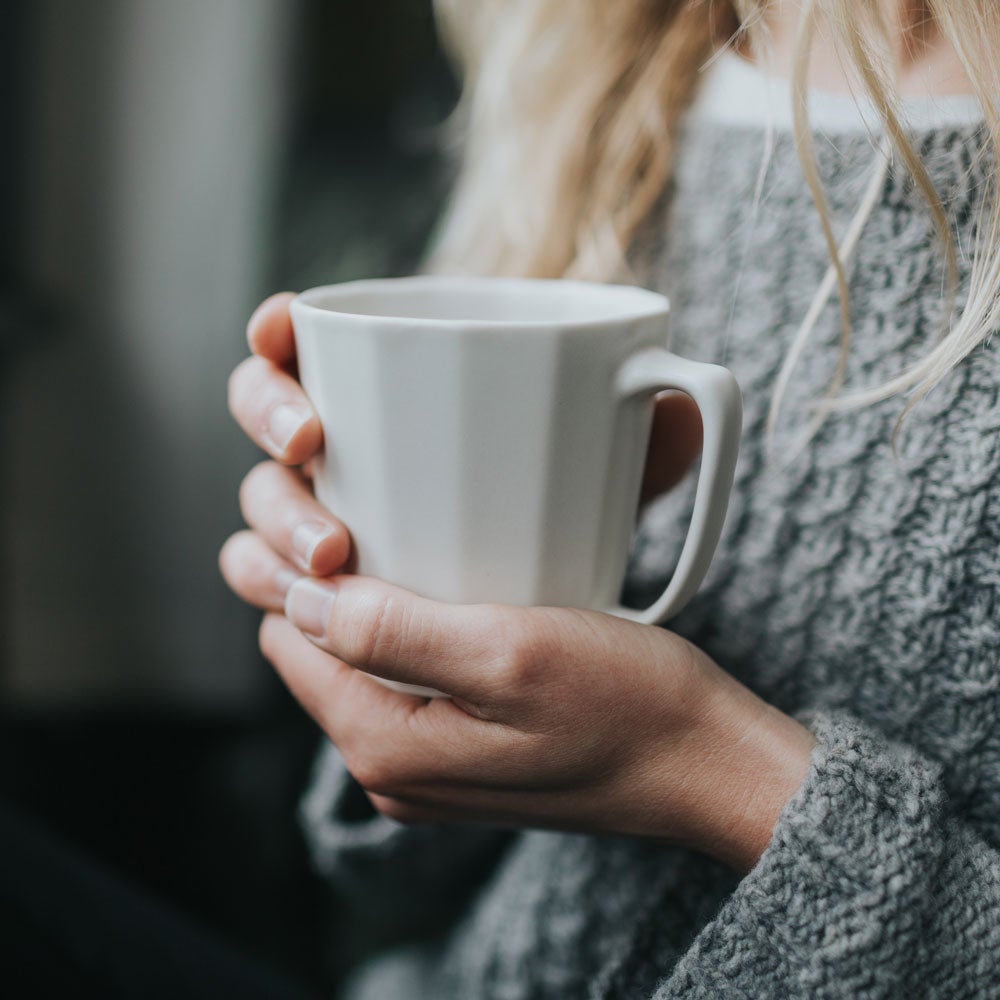Two hands in a grey knit sweater wrapped around a white porcelain coffee mug on a dark background