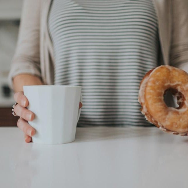 Person in a striped top holding a white porcelain coffee mug at a counter beside a glazed doughnut