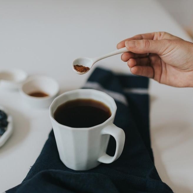 White porcelain mug of black coffee on a dark linen napkin as a hand spoons in sugar from above