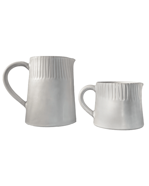 Large and small carved matte white ceramic pitchers side by side on a deep green background