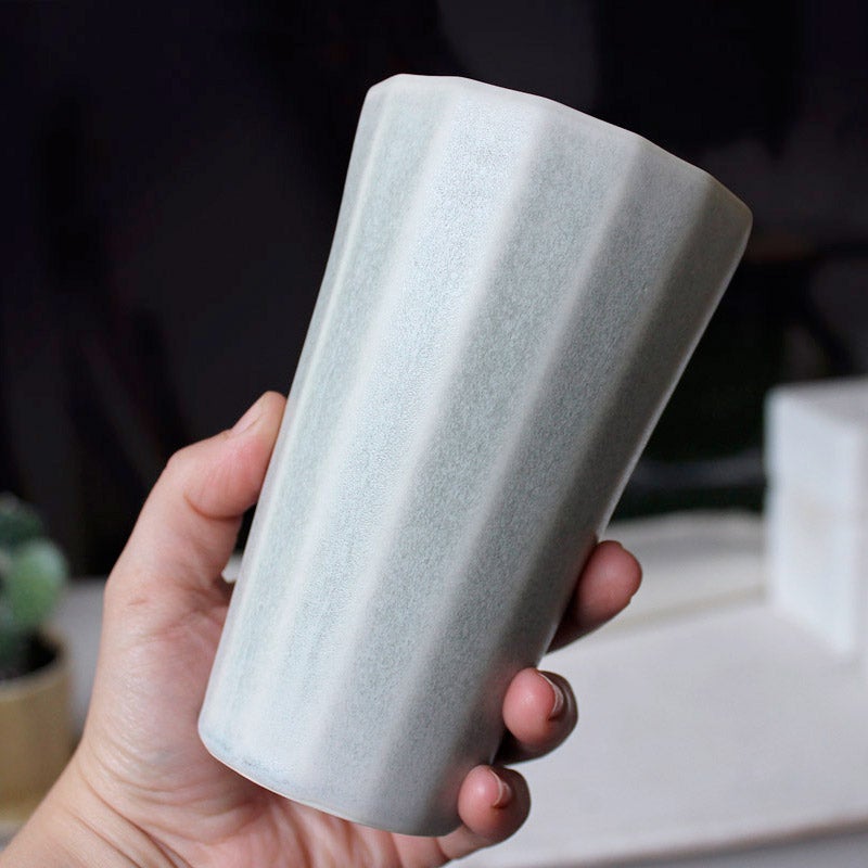 Close view of a hand tilting a pale grey porcelain pint cup to show its faceted sides in soft light
