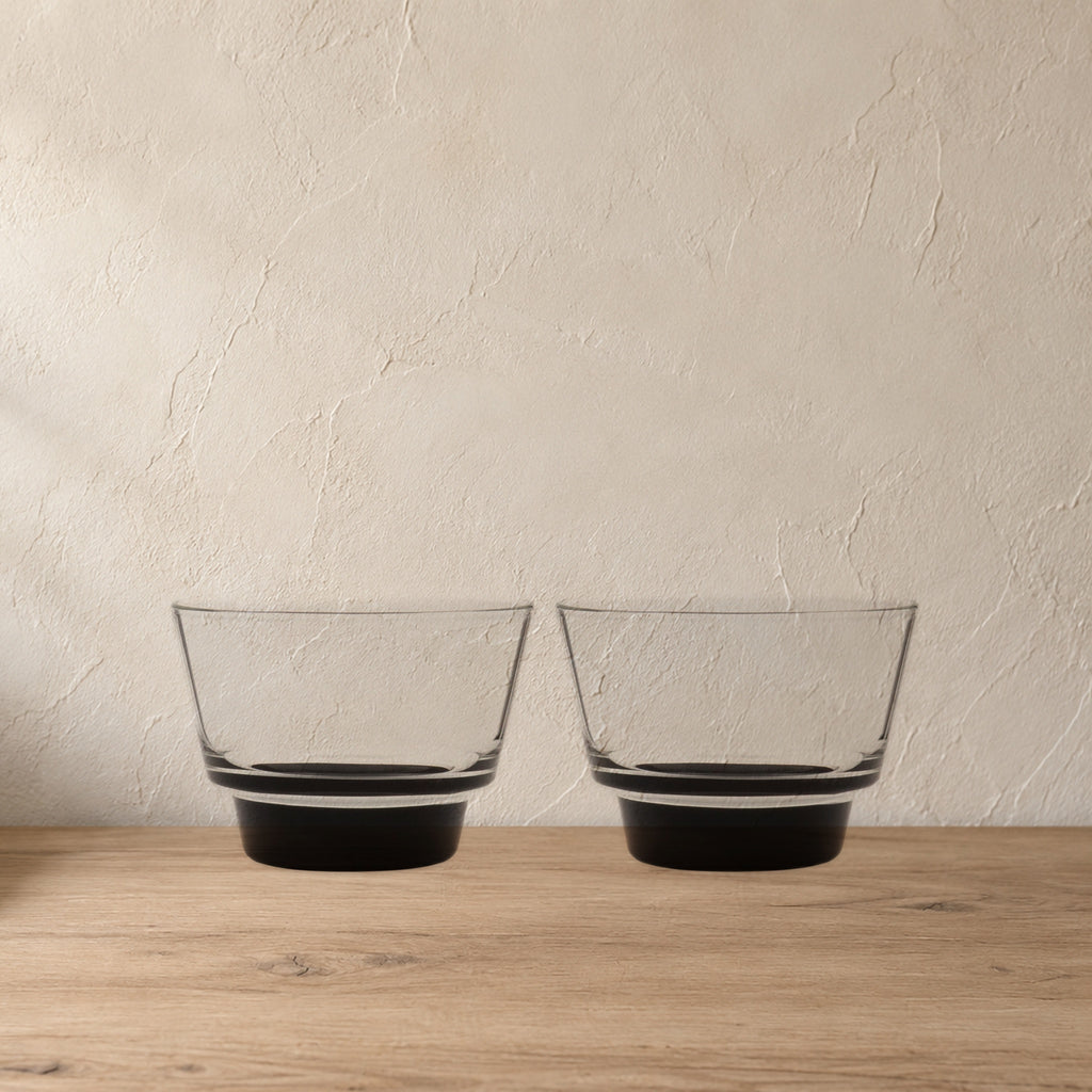 Cirrus Stackable Borosilicate Glass Cups, Black Base, Set of 2 on a pale wood console against a plaster wall.