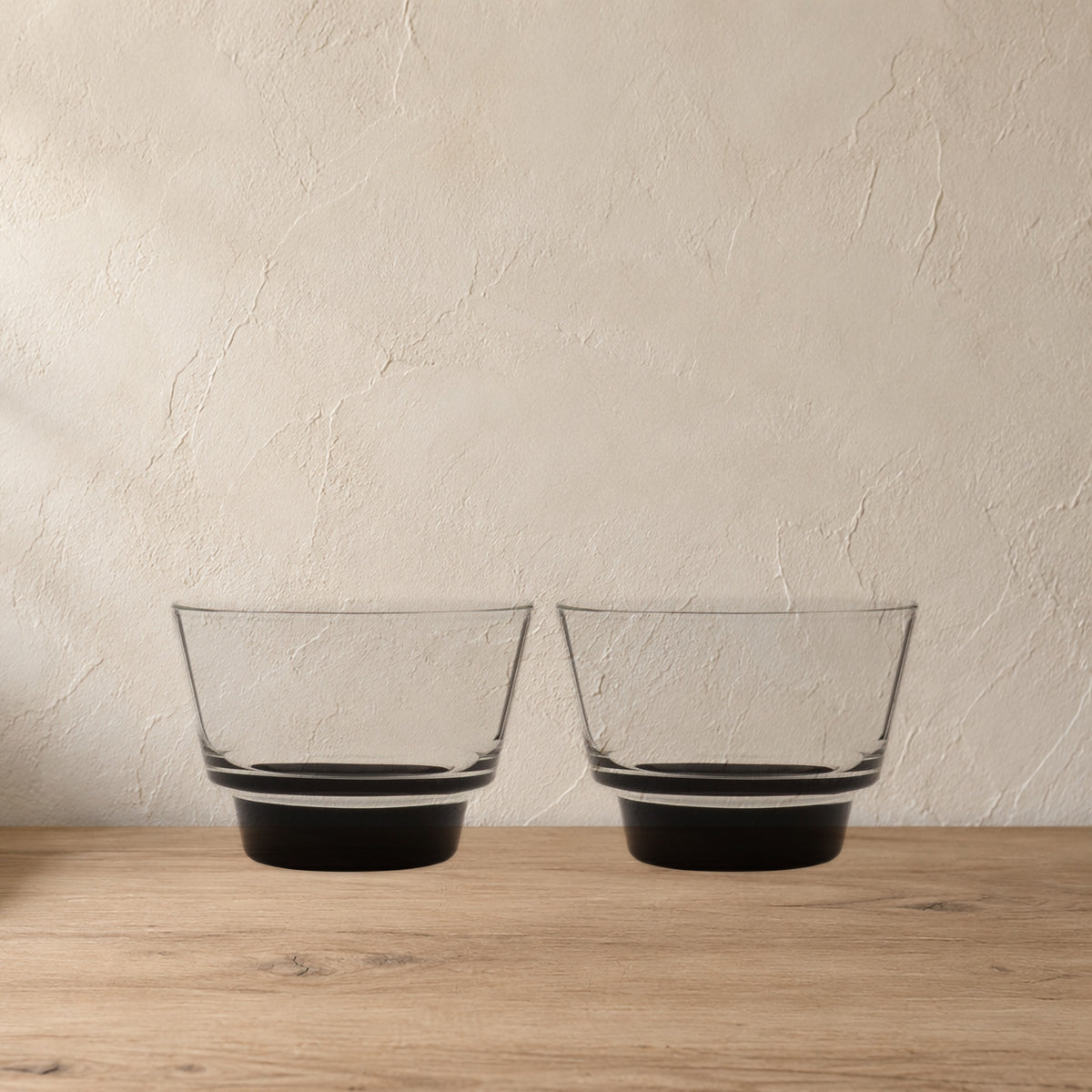 Cirrus Stackable Borosilicate Glass Cups, Black Base, Set of 2 on a pale wood console against a plaster wall.