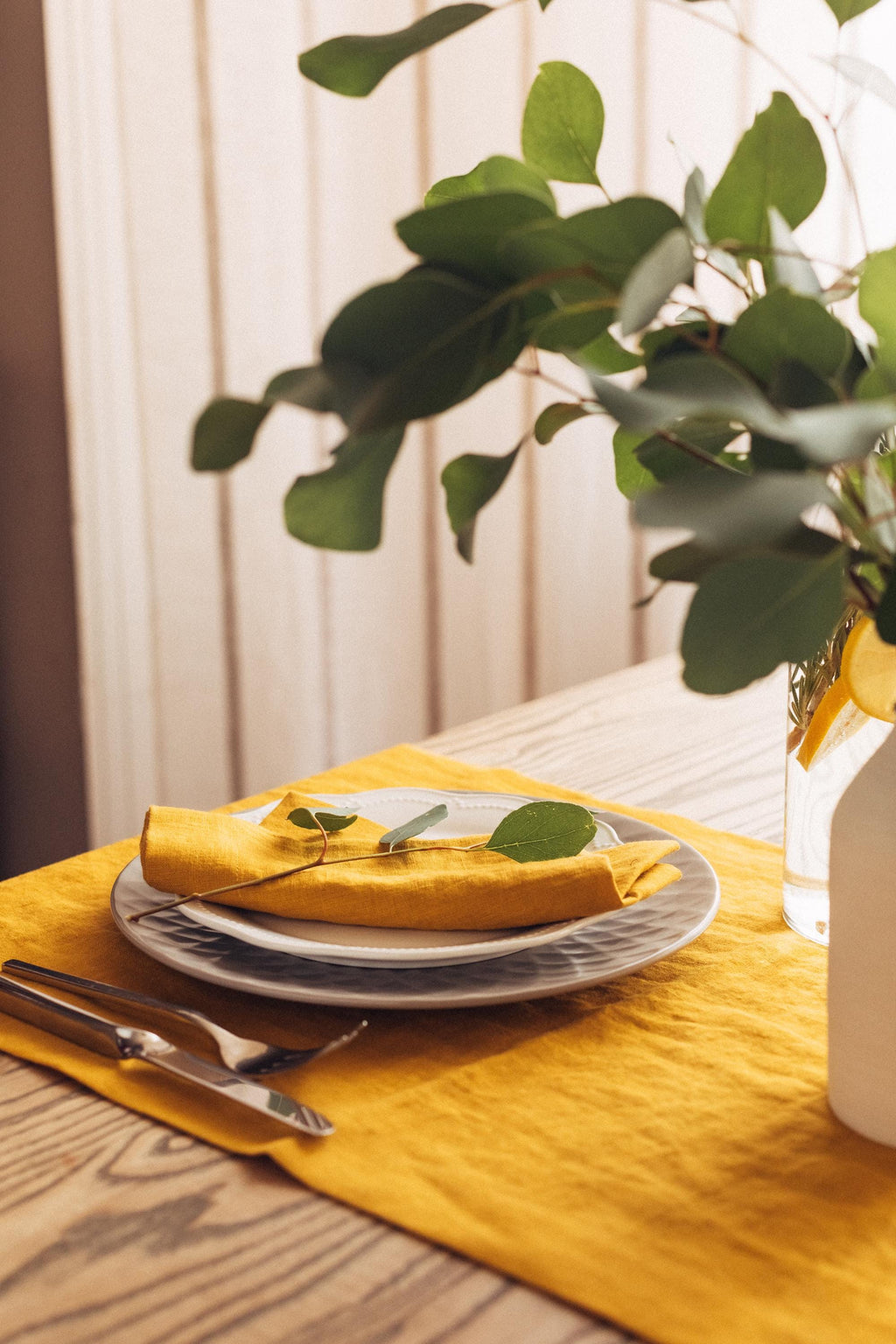 Honey yellow linen runner and napkin on a wood table with layered plates and a vase of green branches